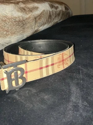 Beiget burberry bälte - Snyggt beige bälte från Burberry med klassiskt rutigt mönster och ett stort metallspänne. Bältet har insida i svart och är tillverkat i skinn. Perfekt accessoar för att lyfta din outfit.