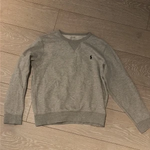 Grå sweatshirt från Polo Ralph Lauren - Klassisk ljusgrå sweatshirt från Polo Ralph Lauren med rund halsringning och den ikoniska lilla broderade loggan på bröstet. Tillverkad i mjukt bomullsmaterial och har ribbade muddar vid ärmslut och nederkant. Perfekt för en avslappnad och stilren look.