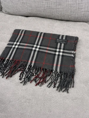 Rutig halsduk från Burberrys i ull - Klassisk halsduk från Burberrys i 100% lammull. Mörkgrå bas med rutigt mönster i vitt, svart och rött. Tidlös design med fransar i ändarna och mjuk känsla. Perfekt accessoar för att lyfta din outfit.