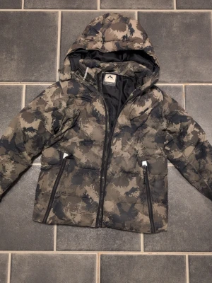 Camomönstrad pufferjacka McKinley - Säljer en camomönstrad pufferjacka från McKinley med huva och två dragkedjeförsedda fickor. Jackan har en cool grå och beige färgskala och är perfekt för kalla vinterdagar. Materialet är vadderat och håller dig varm hela dagen. Finns ett slitage som går att sy lätt, se sista bilden