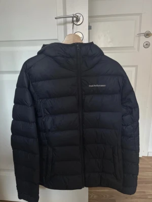 Svart dunjacka Peak Performance - Snygg svart dunjacka från Peak Performance med huva och dragkedja framtill. Jackan har en quiltad design och diskret logga på bröstet. Perfekt för kalla vinterdagar och håller dig varm med sin fluffiga dunfyllning.