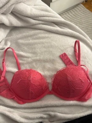 Rosa push-up bh med spetsdetaljer - Säljer en snygg rosa push-up bh med broderade spetsdetaljer på kuporna och justerbara axelband. Bh:n har vadderade kupor och hakknäppning bak. Materialet är en mix av mesh och spets som ger en feminin look.