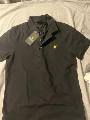 Svart pikétröja från Lyle & Scott - Klassisk svart pikétröja från Lyle & Scott med gul logga på bröstet. Tröjan har krage, knappar framtill och är tillverkad i bomull. Enkel och stilren design som passar till det mesta. Helt ny 