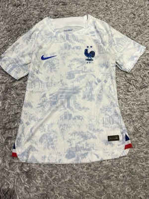 Frankrike fotbollströja Nike blåvit - Säljer en blåvit Frankrike fotbollströja från Nike med diskret mönster av franska landmärken. Tröjan har korta ärmar, blå Nike-logga och landslagets emblem på bröstet. Tillverkad i lätt och ventilerande material, perfekt för träning eller match.