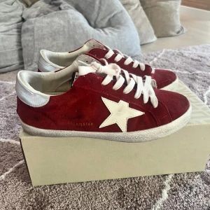 Röda GGDB Superstar sneakers mocka - Snygga röda GGDB Superstar sneakers i mocka med vit stjärndetalj på sidan, vita skosnören och silverfärgad häl. Klassisk platt sula och rund tå. Perfekta för dig som vill sticka ut med en cool streetstyle-look.