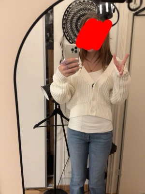 Vit cropped kofta från Kephal - Säljer en vit, stickad cropped kofta från Keepin. Koftan har v-ringning, långa ärmar och stängs med två knappar framtill. Den är ribbstickad och har en avslappnad passform som passar perfekt till jeans eller kjol.