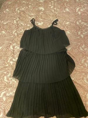 Svart plisserad långklänning - Svart långklänning med tunna axelband och knyt på axlarna. Klänningen har plisserade lager och ett luftigt, flowy fall. Perfekt för dig som gillar en elegant och enkel stil.