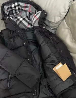 Svart pufferjacka från Burberry - Snygg svart pufferjacka från Burberry med klassiskt rutigt foder i huvan. Jackan har dragkedja och knappar framtill, flera fickor och en hög krage. Materialet är polyester med dunfyllning för extra värme. Perfekt för kalla vinterdagar.