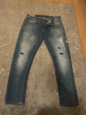 Dondup jeans med slitningar  - Säljer ett par blå dondup jeans med slitningar och hål på benen för en riktigt avslappnad och trendig look. Jeansen har klassisk femficksmodell, slimmade ben och detaljerade slitningar framtill. Perfekta för dig som gillar streetstyle och vill ha en cool vibe.