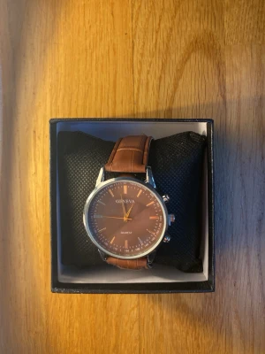 Brun klocka från Geneva med läderband - Stilren klocka från Geneva med brunt läderband och rund urtavla i matchande brun ton. Urtavlan har silverfärgade detaljer och tydliga markeringar. Klockan har klassisk design och passar perfekt till alla outfits.