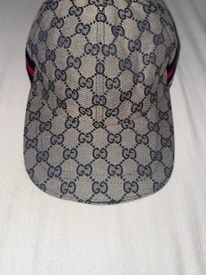 Gucci beige keps med GG-mönster - Säljer en beige keps från Gucci med det klassiska GG-monogrammet över hela kepsen. På sidan finns en snygg detalj med blå och röd rand. Kepsen har böjd skärm och är tillverkad i canvasmaterial som ger en exklusiv känsla.