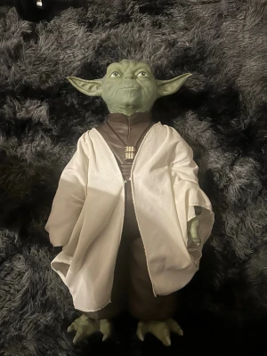 Yoda maskeradaccessoar Star Wars - Maskeradaccessoar föreställande Yoda från Star Wars. Grön mask med stora öron, bruna kläder och vit mantel i tyg. Perfekt för cosplay eller temafest. Detaljerad design med autentiskt utseende och karaktäristiska drag. Köpt 2017. Samlar figur-ej leksak. Priset kan diskuteras