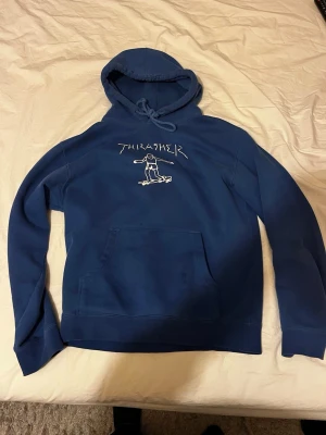 Blå Thrasher hoodie med tryck - Säljer en blå hoodie från Thrasher med vit tryckt logga och en skatare på bröstet. Klassisk känguruficka framtill, dragsko i huvan och mjukt material. Perfekt för dig som gillar streetwear och skatekultur.