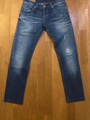 Blå slitna jeans från märket Dondup - Säljer ett par blå jeans från märket dondup med snyggt slitna detaljer och patch på höger ben. Jeansen har raka ben, klassisk femficksmodell och kontrastsömmar. Perfekta för en avslappnad och trendig look.