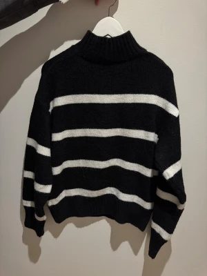 Svart stickad tröja med vita ränder - En svart stickad tröja med vita horisontella ränder och hög krage. Tröjan har en avslappnad passform, långa ärmar och ribbstickade muddar vid hals, ärmslut och nederkant. 