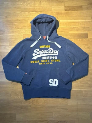 Superdry hoodie - Säljer en blå hoodie från Superdry med vit och gul text på bröstet. Storlek L och perfekt skick. Hör av dig vid frågor eller prisförslag!