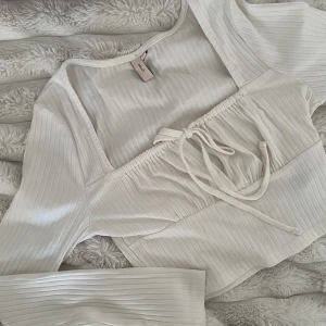 Vit ribbad croppad topp med snörning från NELLY - Trendig vit croppad topp från NELLY med ribbad struktur och långa ärmar. Toppen har snygg snörning framtill och en lätt v-ringad halsringning. Perfekt för att skapa en stilren och modern look.