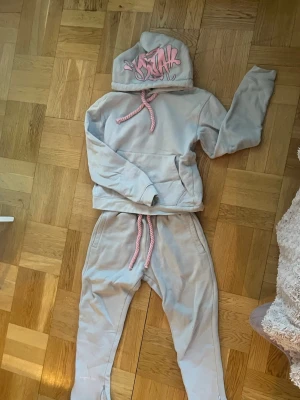 Ljusgrå mjukisbyxor Stay med rosa snören - Säljer ett par ljusgrå mjukisbyxor från Syna World med rosa dragsko i midjan och dragkedjor vid bensluten. Byxorna har fickor på sidorna och är tillverkade i mjukt bomullsmaterial. Perfekta för chill dagar och streetwear-stil.