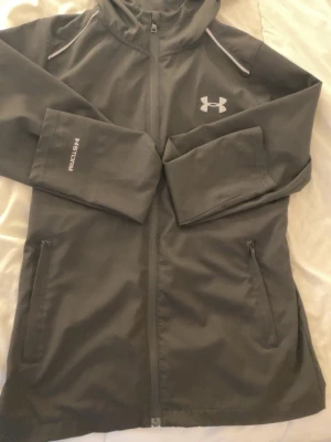 Svart vindjacka från Under Armour - Snygg svart vindjacka från Under Armour med huva och hel dragkedja. Jackan har två fickor med dragkedja, vita detaljer på axlarna och logga fram samt stor logga på ryggen. Tillverkad i lätt och vattenavvisande material med UA Storm-teknologi.