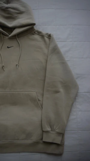 Nike Logo Hoodie  - Snygg beige hoodie från Nike med klassisk svart swoosh-logga broderad på bröstet. Hoodien har en stor känguruficka framtill, ribbade muddar vid ärmslut och nederkant samt justerbar huva med dragsko. Perfekt för en avslappnad och sportig stil. Limited edition och stilren för gymmet! 