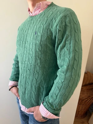 Grön kabelstickad tröja Polo Ralph Lauren - Kabelstickad grön tröja från Polo Ralph Lauren med rund halsringning och diskret broderad logga på bröstet. Storlek L i bra skick, skjortan under ligger också ute på min plick!