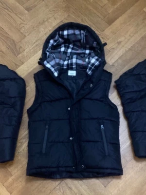 Svart dunväst med avtagbara ärmar - Svart dunväst med avtagbara ärmar och huva. Fodret har rutigt mönster i svart, vitt och grått. Två dragkedjefickor framtill och stängning med både dragkedja och knappar. Perfekt för lager-på-lager och streetstyle.