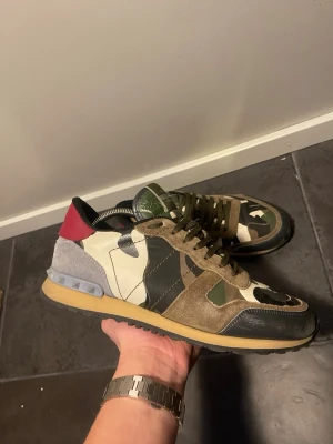Valentino camo sneakers med mockadetaljer - Säljer ett par unika Valentino sneakers med camomönster i grönt, beige och svart. Skorna har detaljer i mocka, läder och textil samt en röd hälflik. Snygg kontrast mellan material och färger, perfekt för dig som vill sticka ut.