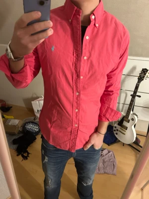 Rosa skjorta från Ralph Lauren - Säljer en snygg rosa skjorta från Ralph Lauren med klassisk button-down krage och det ikoniska broderade logotypen på bröstet. Skjortan har lång ärm, knappar framtill och en normal passform. Perfekt för dig som vill ha en stilren och tidlös look.