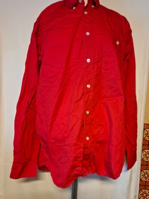 Röd skjorta med knappar - Klassisk röd skjorta med lång ärm och vita knappar framtill. Skjortan har en enkel design med krage och är tillverkad i bomull, perfekt för en stilren look. Passar dig som gillar färg och vill sticka ut lite extra. Tyvärr så är inte originalförpackningen kvar för den var trasig. Tvättad på grund av att förpackningen var trasig. Finns heller ingen prisetikett kvar. Skjortan är aldrig använd. 