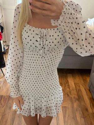 Vit prickig kortklänning med volang - Supersöt vit kortklänning med svarta prickar, långa puffiga ärmar och volangkant nertill. Klänningen har smockad midja, knyt i ringningen och är tillverkad i ett lätt, luftigt material som känns perfekt för varma dagar.