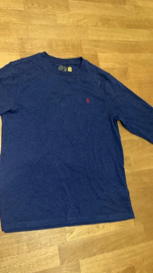 Blå långärmad topp från Polo Ralph Lauren - Mörkblå långärmad topp från Polo Ralph Lauren med rund halsringning och diskret röd logga broderad på bröstet. Tillverkad i mjuk bomull, perfekt för en avslappnad och stilren look.