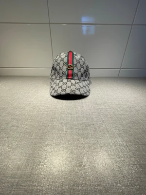 Gucci keps med GG-mönster - Säljer en snygg keps från Gucci med klassiskt GG-mönster i grått och svart. Kepsen har en röd och grön rand längs mitten och ett guldfärgat GG-emblem framtill. Justerbar baktill och tillverkad i ett slitstarkt tygmaterial.