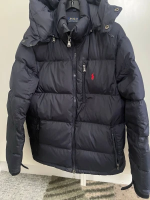 Svart dunjacka från Polo Ralph Lauren - Snygg svart dunjacka från Polo Ralph Lauren med röd logga på bröstet. Jackan har huva, flera dragkedjeförsedda fickor och en hög krage. Perfekt för kalla vinterdagar och riktigt skön att bära. Klassisk puffer-modell med stilrena detaljer.