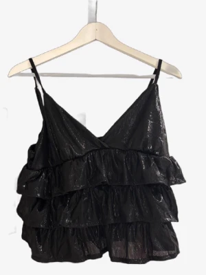 Svart glittrig volangtop - Snygg svart glittrig topp med tunna axelband och flera lager volanger. Toppen har omlott-look framtill och är perfekt för dig som vill sticka ut lite extra. Passar till många olika stilar och är superenkel att matcha.