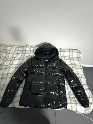 Svart pufferjacka från Trapstar - Säljer en svart, glansig pufferjacka från Trapstar med huva och dragkedja. Jackan har broderad logga på bröstet och stor Trapstar-text på ryggen. Tillverkad i 100% polyester och har en loose passform som ger en chill vibe. Perfekt för kalla dagar.