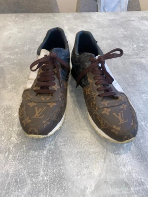 Louis Vuitton bruna sneakers - Snygga sneakers från Louis Vuitton med klassiskt monogrammönster i brunt och beige. Skorna har bruna skosnören, vita detaljer på sidorna och svart insida. Tillverkade i läder med LV-logga över hela ovandelen. Perfekta för dig som vill sticka ut med lyxig streetstyle.
