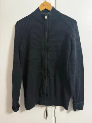 Svart stickad ziptröja med struktur - Snygg svart stickad ziptröja med hög krage och dragkedja framtill. Tröjan har ett coolt strukturerat mönster över bröst, rygg och ärmar samt ribbade muddar. Perfekt för dig som gillar stilrena och moderna plagg.