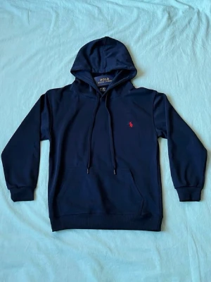 Mörkblå hoodie från Polo Ralph Lauren skick 10/10 aldrig använd  - Snygg mörkblå hoodie från Polo Ralph Lauren med klassisk huva och dragsnören. Har en röd broderad logga på bröstet och stor magficka framtill. Tillverkad i mjukt bomullsmaterial som känns skönt mot huden. Perfekt för en avslappnad och stilren look. Stl s men passar xs, 165