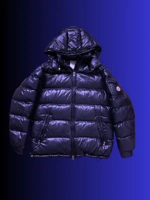 Moncler Maya Jacka - ÄKTA:  Snygg svart dunjacka från Moncler med glansig finish och stor huva. Jackan har två dragkedjefickor fram, Moncler-logga på ärmen och breda kanaler fyllda med dun. Perfekt för kalla vinterdagar och riktigt trendig look.