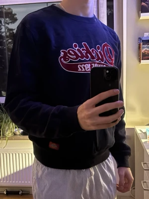 Marinblå Dickies sweatshirt med tryck - Säljer en marinblå sweatshirt från Dickies med stort rött och vitt tryck på bröstet där det står 'Dickies Since 1922'. Tröjan har rund halsringning, ribbade muddar och en klassisk passform. Perfekt för en avslappnad streetwear-look.