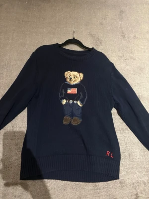 Mörkblå stickad tröja  - Snygg mörkblå stickad tröja från Ralph Lauren med ikoniskt björnmotiv på bröstet och broderade initialer 'RL' i rött vid nederkanten. Tröjan har rund halsringning, långa ärmar och ribbstickade muddar. Perfekt för dig som gillar klassiska och lekfulla detaljer.