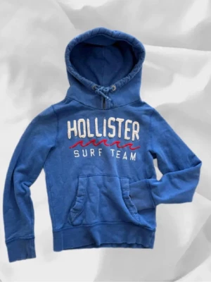 Blå Hollister hoodie med tryck - Snygg blå hoodie från Hollister med stor huva och dragsko. Framtill står det 'HOLLISTER SURF TEAM' i vitt och rött tryck. Hoodien har en klassisk känguruficka och ribbade muddar vid ärmar och nederkant. Perfekt för en avslappnad och sportig stil.