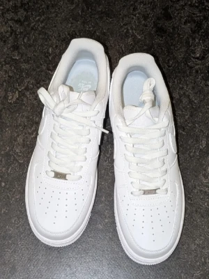 Nike Air Force 1 - Klassiska helvita Nike Air Force 1 sneakers i läder med låg profil och tydlig swoosh-logga på sidorna. Skorna har tjock sula, snörning och diskret Nike Air-märkning på hälen. Perfekta för en clean och stilren look.