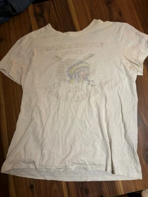 Vit t-shirt från Denim & Supply Ralph Lauren - Vit t-shirt från Denim & Supply Ralph Lauren med tryck av ett huvudbonadsmotiv och text på bröstet. Klassisk rund hals och korta ärmar. Tillverkad i mjuk bomull för en skön känsla och avslappnad look.