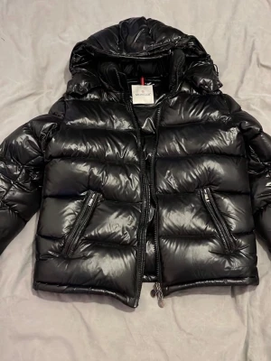 Svart dunjacka från Moncler - Snygg svart dunjacka från Moncler med glansig finish, huva och två dragkedjeförsedda fickor framtill. Jackan har quiltad design och dragkedja hela vägen. Perfekt för kalla vinterdagar och riktigt trendig look. Ett spänne är sönder på baksidan, vid midjan!.