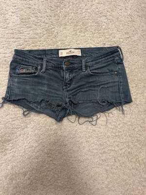 Shorts - Snygga blå jeansshorts från Hollister med råa, fransiga kanter och klassisk femficksdesign. Shortsen har en väldigt låg midja och slitna detaljer för en avslappnad look. Perfekta för varma dagar och sommarhäng. 