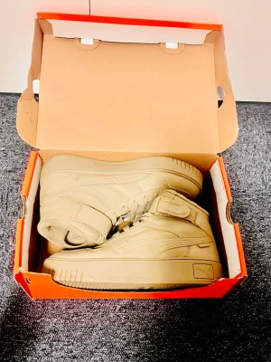 Sneakers Puma - Snygga beige sneakers från Puma i högt modell med kardborreband och klassisk logga på sidan. Skorna är tillverkade i läder och har matchande beige sula och snörning. Perfekta för dig som vill ha en clean och stilren look.
