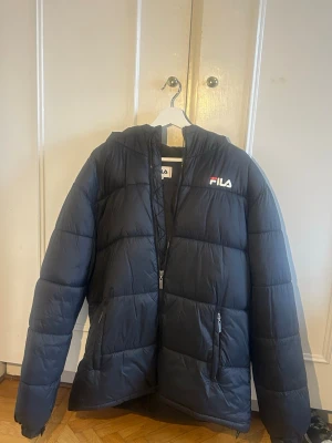 Svart pufferjacka från FILA - Snygg svart pufferjacka från FILA med stor logga på ryggen och mindre logga på bröstet. Jackan har huva, dragkedja framtill och två sidofickor med dragkedja. Perfekt för kalla dagar och ger en sportig vibe. Materialet är syntetiskt och jackan är quiltad för extra värme.