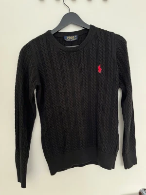 Svart kabelstickad tröja Polo Ralph Lauren - Kabelstickad svart tröja från Polo Ralph Lauren med rund halsringning och röd broderad logga på bröstet. Långa ärmar och ribbade muddar vid ärmslut och nederkant. Klassisk och stilren design som passar till det mesta.