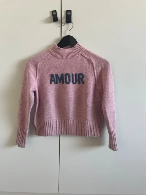 Rosa stickad tröja Zadig & Voltaire - Snygg rosa stickad tröja från Zadig & Voltaire med texten 'AMOUR' i mörkblått framtill. Tröjan har ribbade muddar vid hals, ärmslut och nederkant samt en avslappnad passform. Perfekt för dig som gillar trendiga och bekväma plagg.